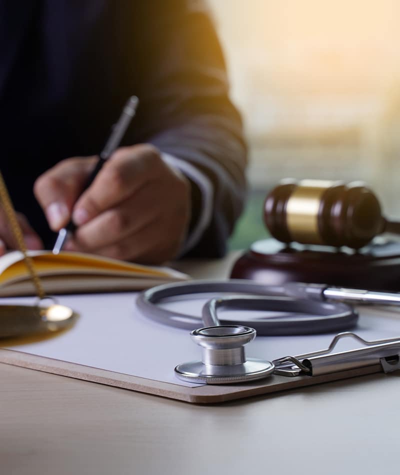 A stethoscope and gavel sitting on a clipboard featuring a hand writing in the background.