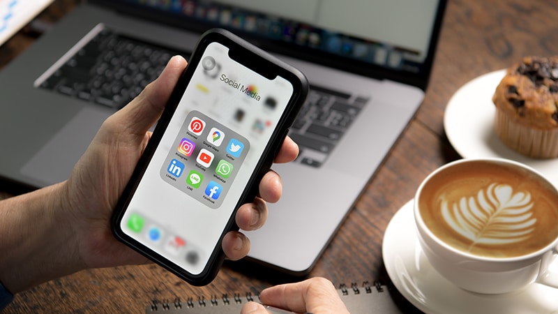 A hand holding an iPhone showing various social media apps.